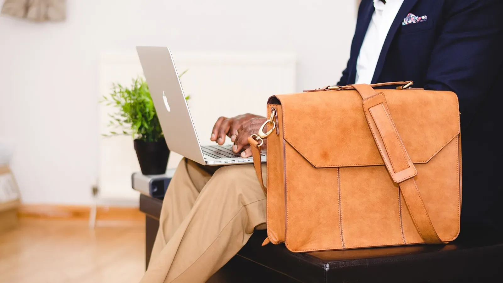 Pria berjas mengetik di laptop sambil duduk dengan tas kulit di sampingnya